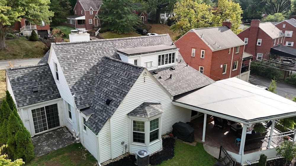 White house with grey roof and attached carport. Red brick building in the background, green trees surround.