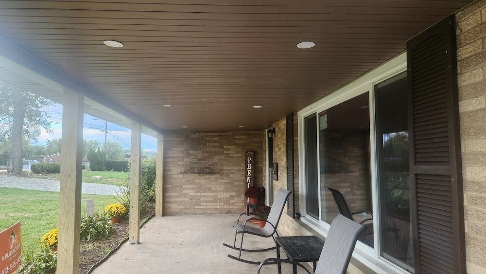Covered porch with brown ceiling and brick wall. Sliding glass windows with black shutters. Black chairs sit on the porch.