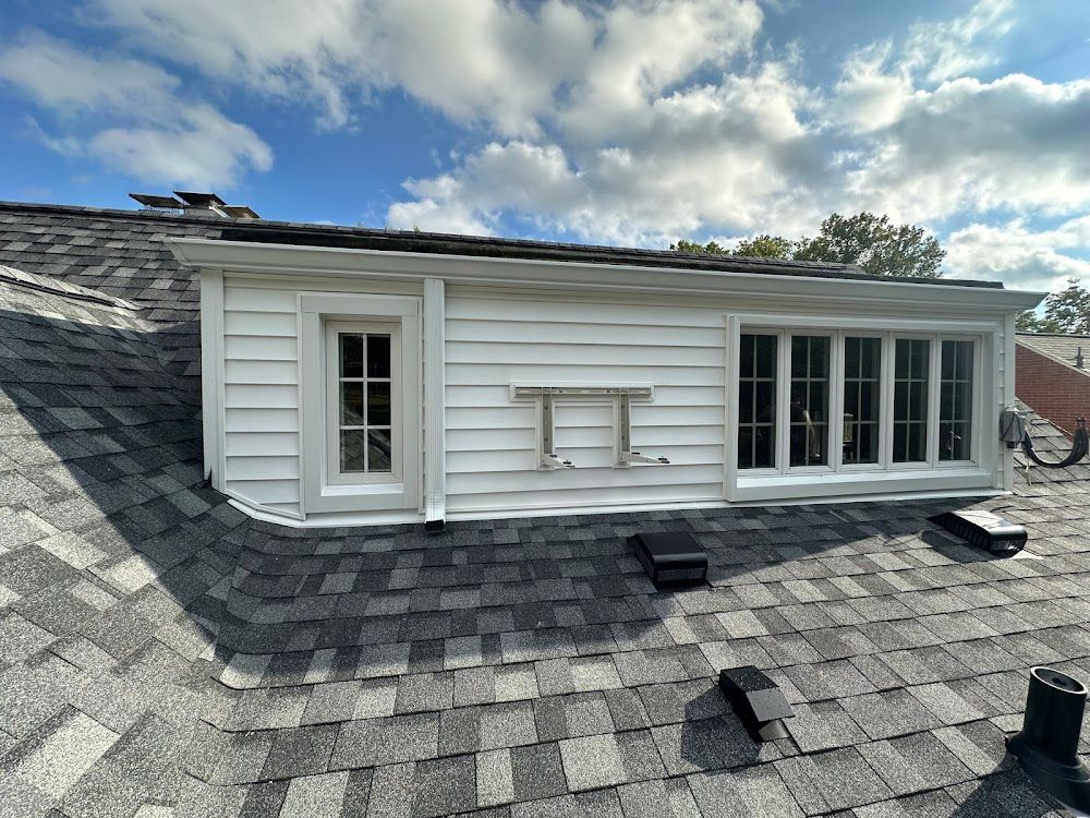 White dormer with windows on a gray shingled roof, set against a partly cloudy blue sky.