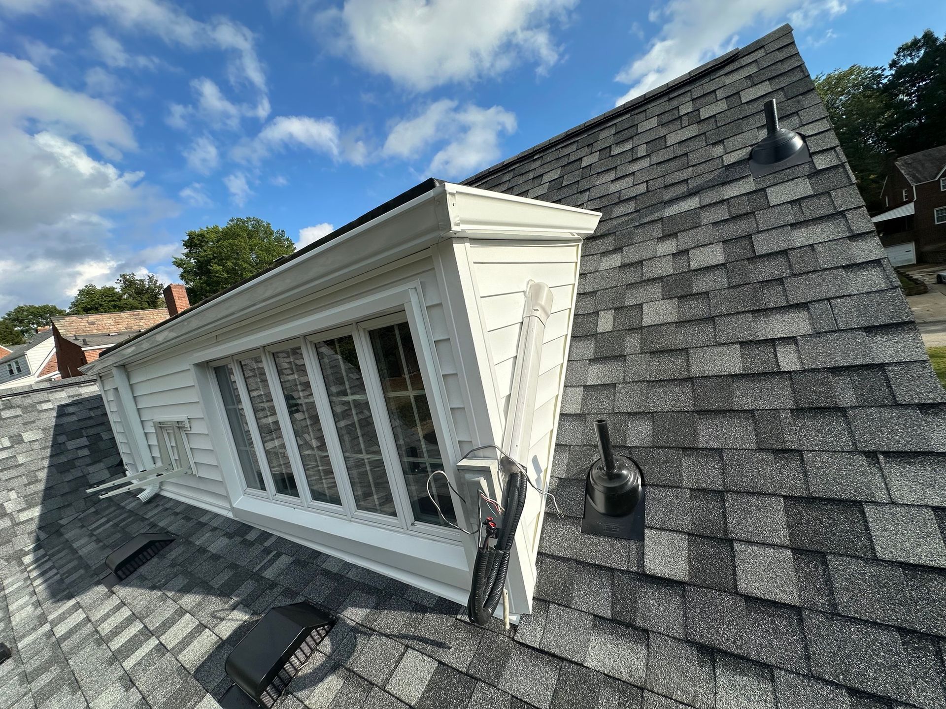 A roof with gray shingles and a white dormer window under a blue sky with clouds.