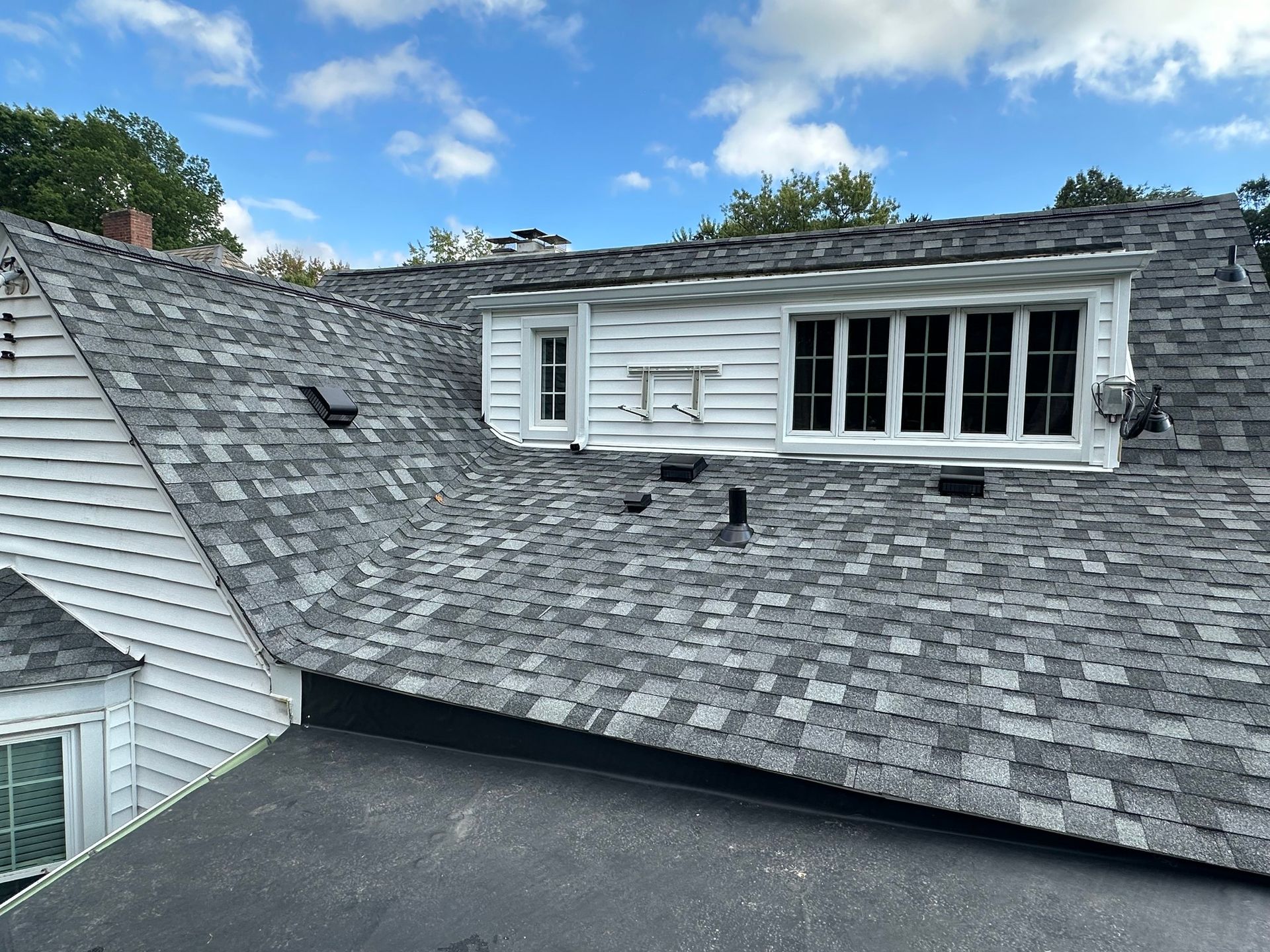 Gray shingle roof of a house with a white dormer window and blue sky background.