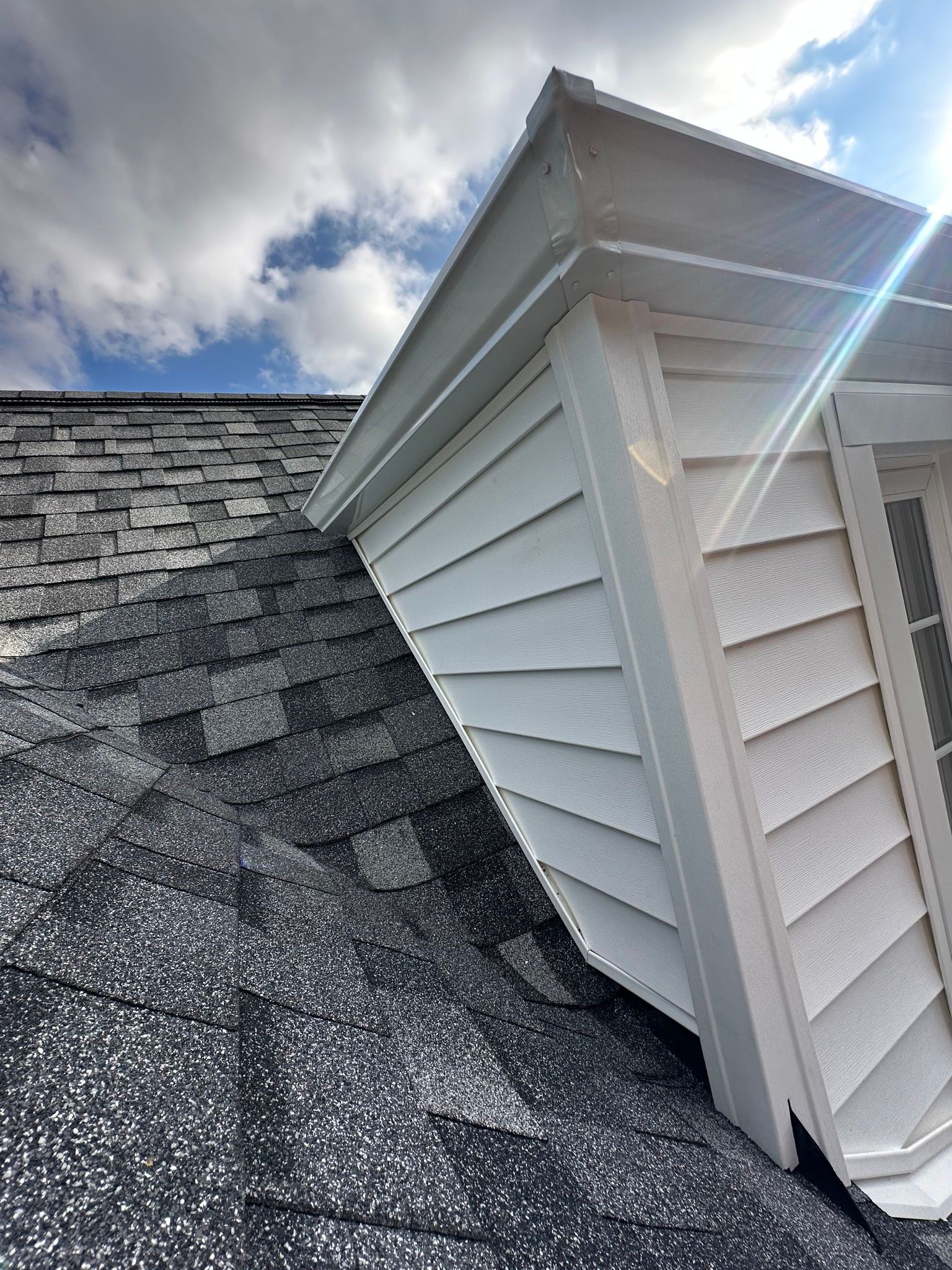 Close-up of a white dormer window on a roof with dark gray shingles against a cloudy sky.