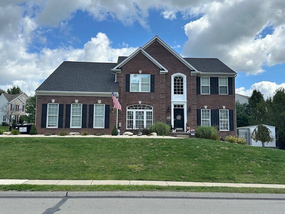 Two-story brick house with black shutters, American flag, and manicured lawn under a cloudy blue sky.