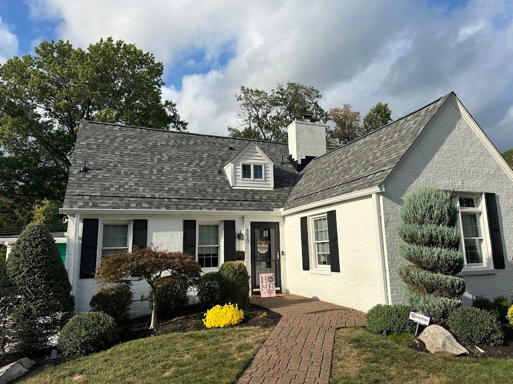 White house with a gray roof and black shutters, brick path leading to the door, bushes and trees in the yard.