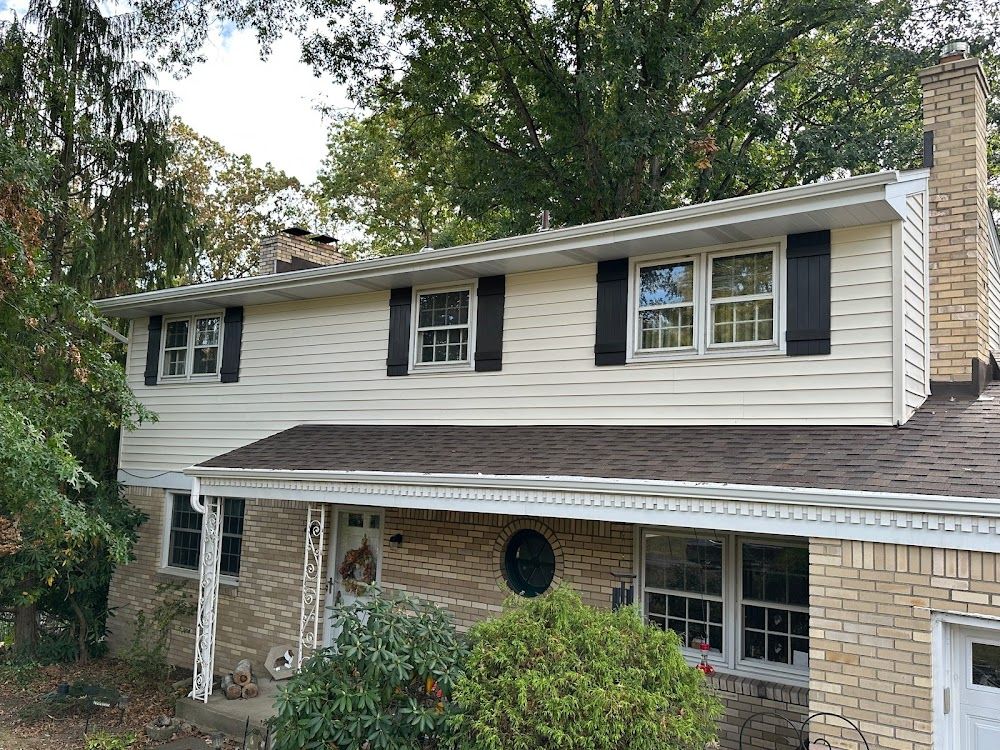 Beige two-story house with black shutters, brown roof, and a brick chimney surrounded by trees.