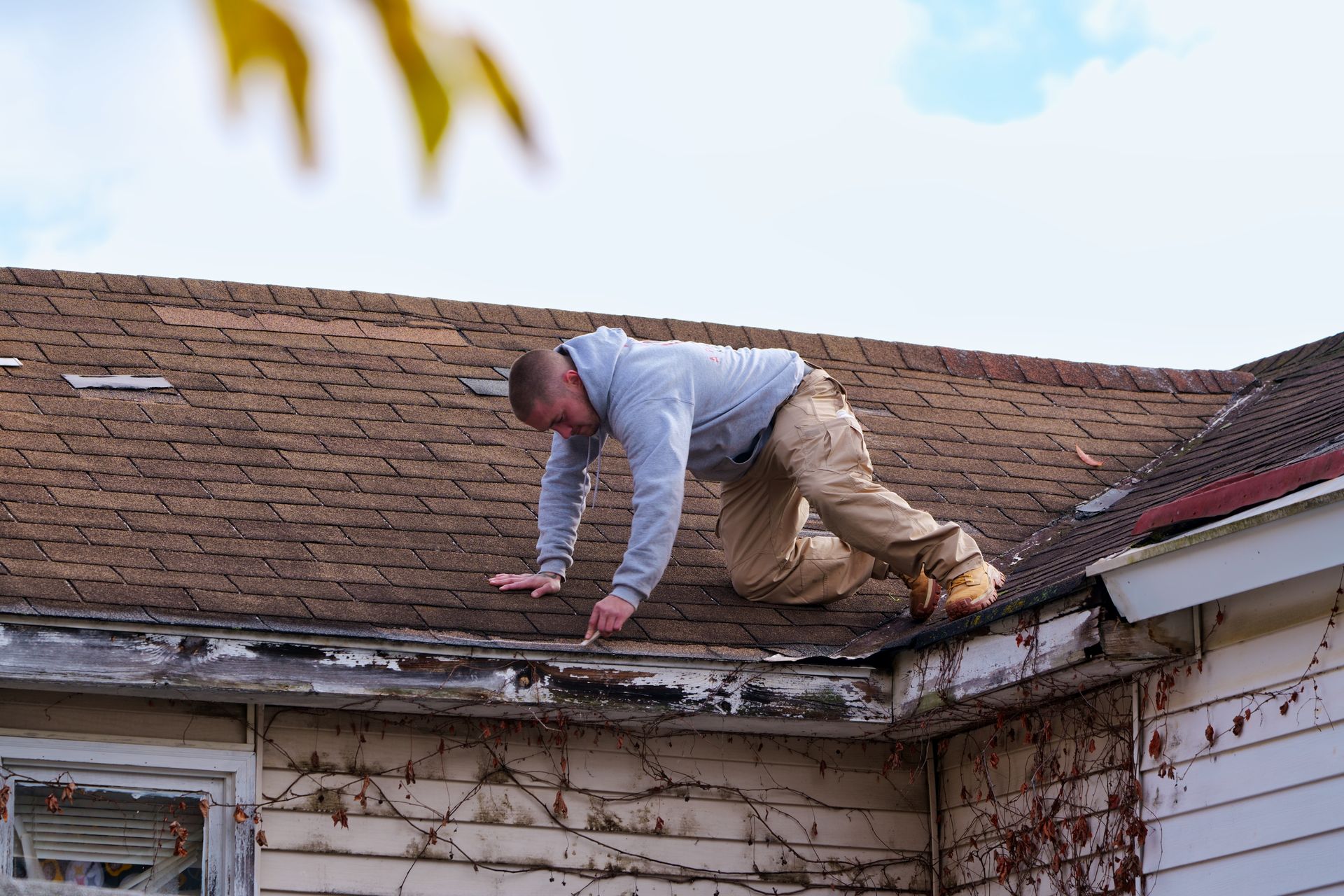 Man on a roof, inspecting the edge. Overcast sky. The house has peeling paint and vines growing on it.