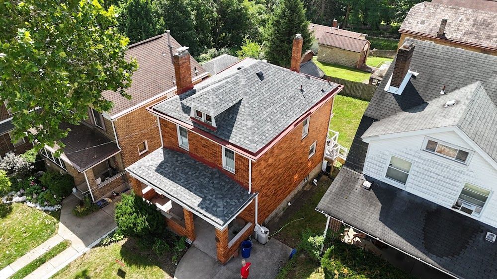 Aerial view of brick house with dark gray roof, red trim, and surrounding houses with green trees.