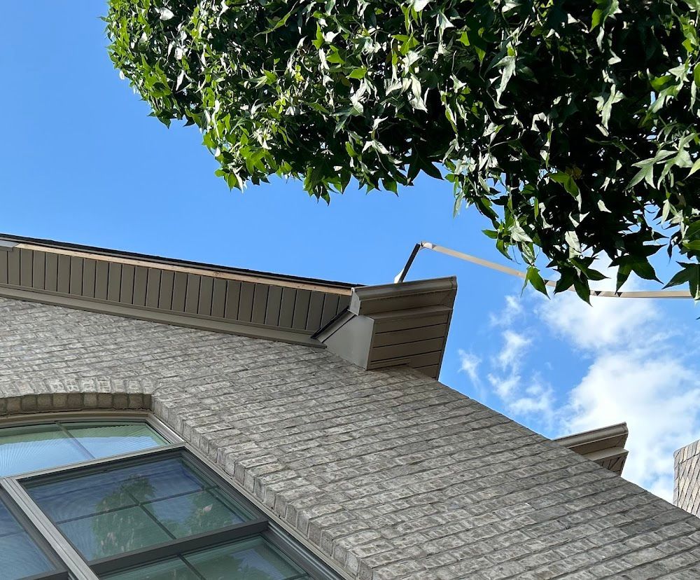 Brick building with a window, roof, and leafy tree against a bright blue sky.