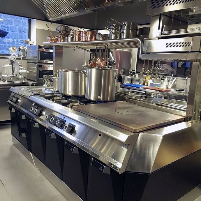 Cucina professionale in acciaio inox con forni, frigoriferi e tavoli da lavoro illuminati da luci intense.