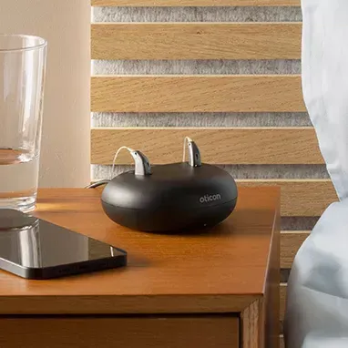 Black wireless earbuds case on a wooden nightstand beside a phone and glass, with a striped headboard behind.