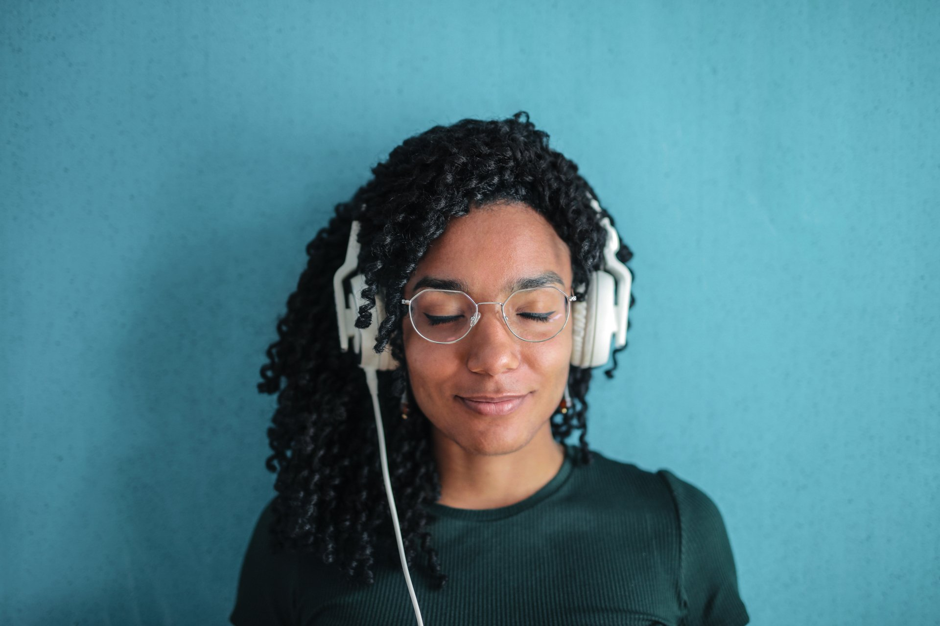 Person with curly hair and glasses wearing headphones against a teal background, eyes closed.