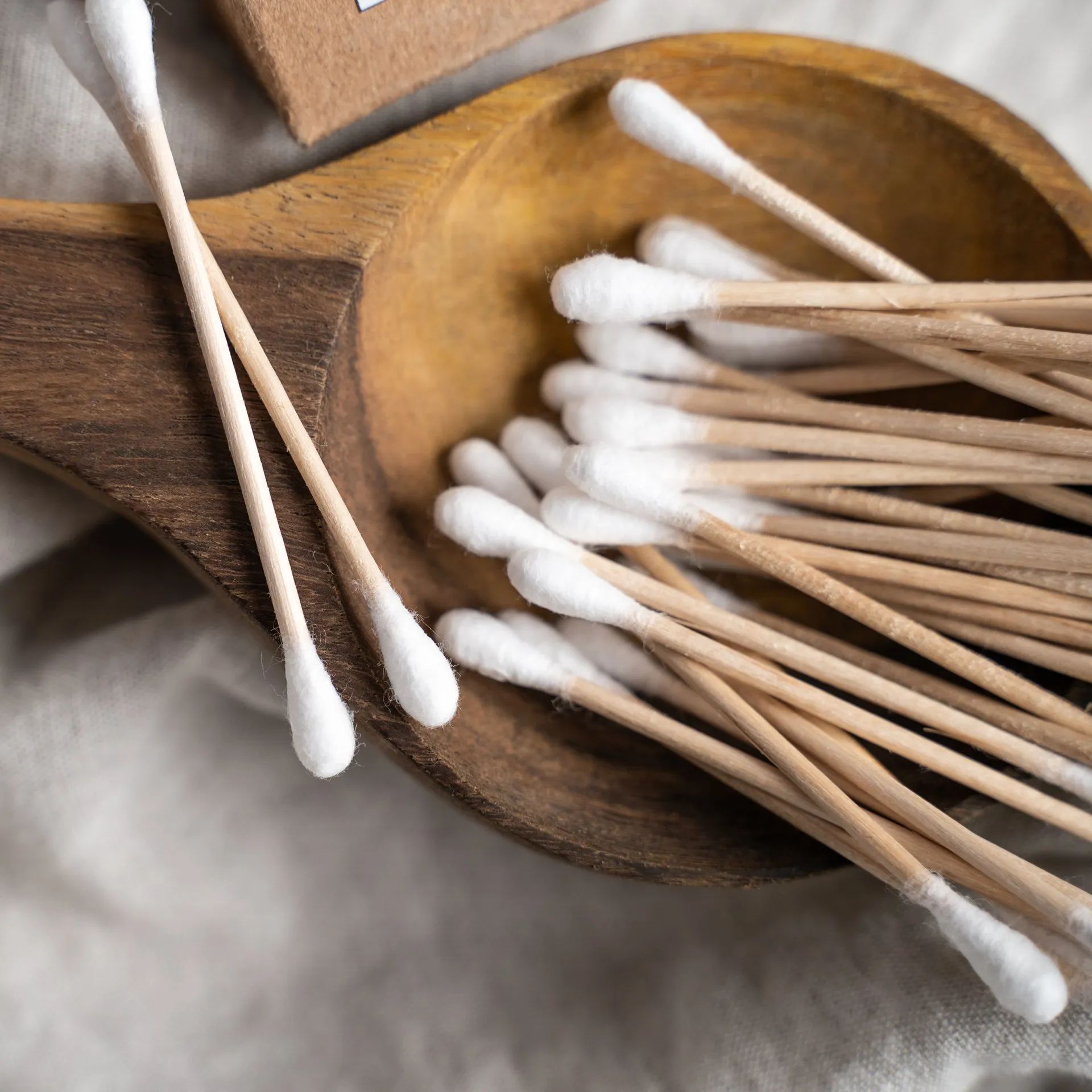 Cotton swabs in a wooden bowl and scattered on a soft beige fabric background