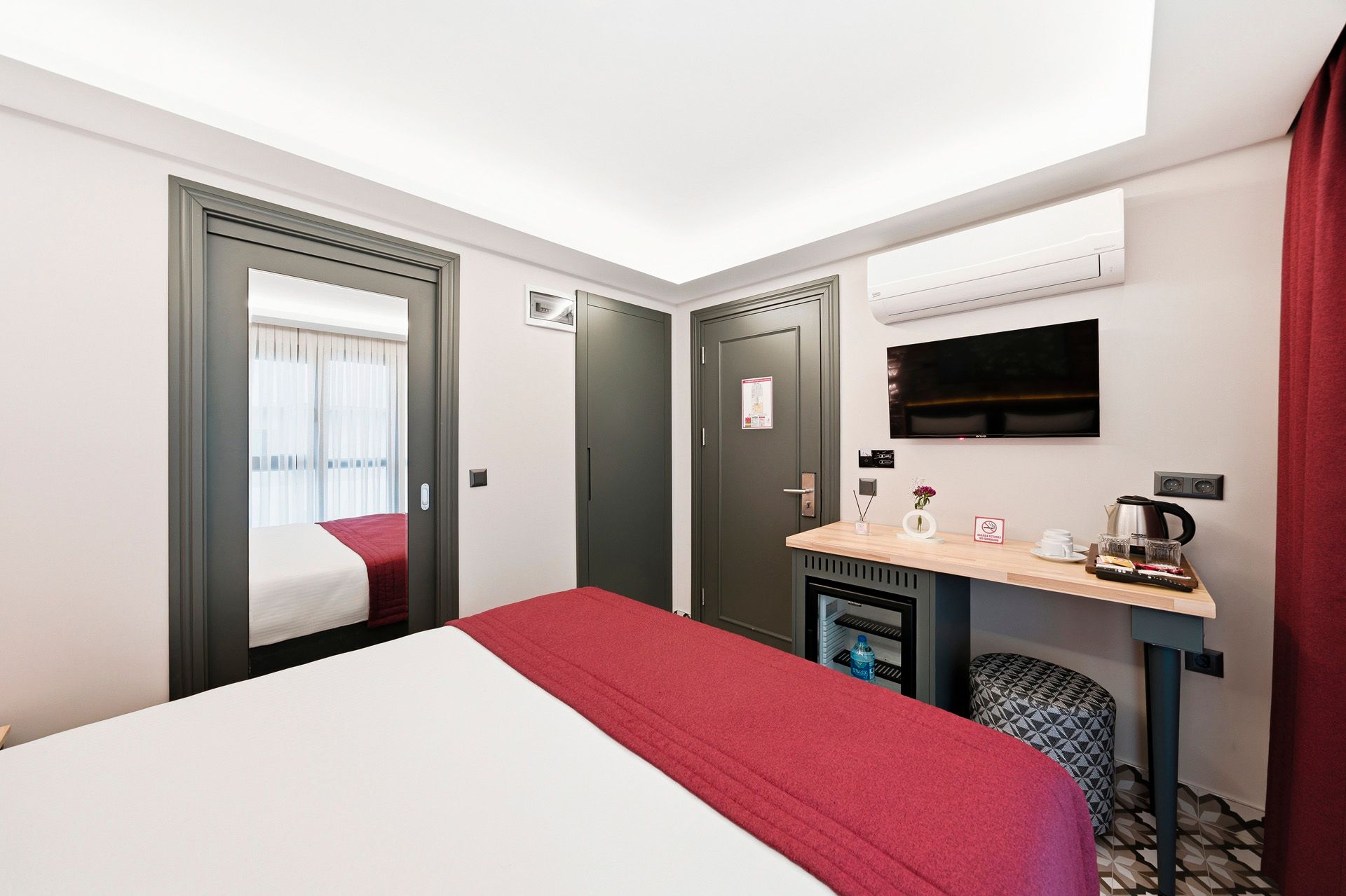 Modern hotel room with bed, wall-mounted TV, desk, mirror, and red accents