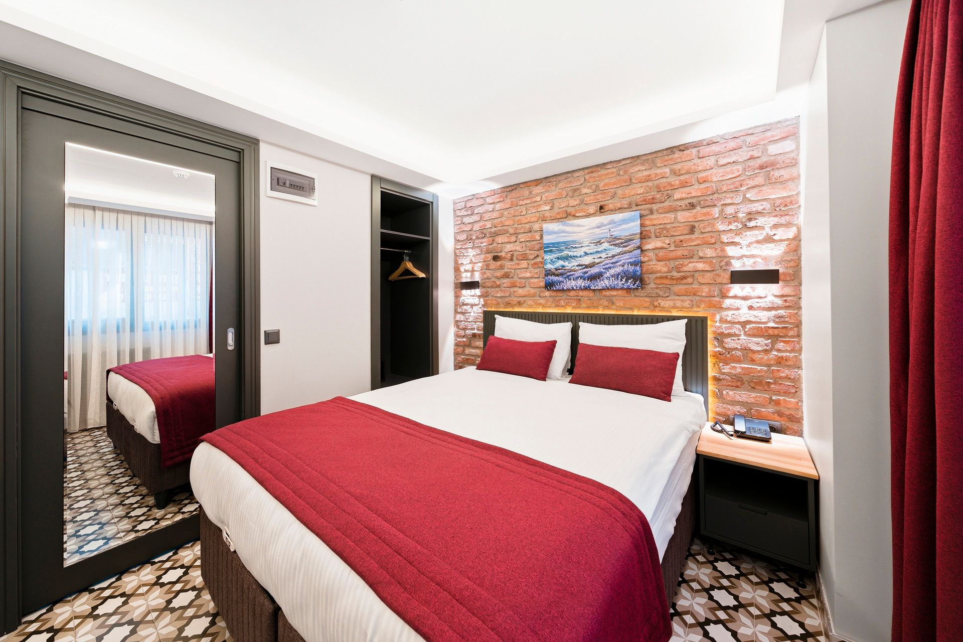 ARTELIST HOTEL | Modern hotel bedroom with a red bedspread, brick accent wall, mirror closet, and window.