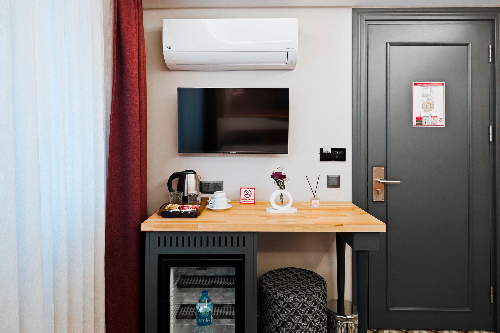 ARTELIST HOTEL | Small hotel room corner with wall-mounted TV, air conditioner, desk, mini fridge, chair, and gray door