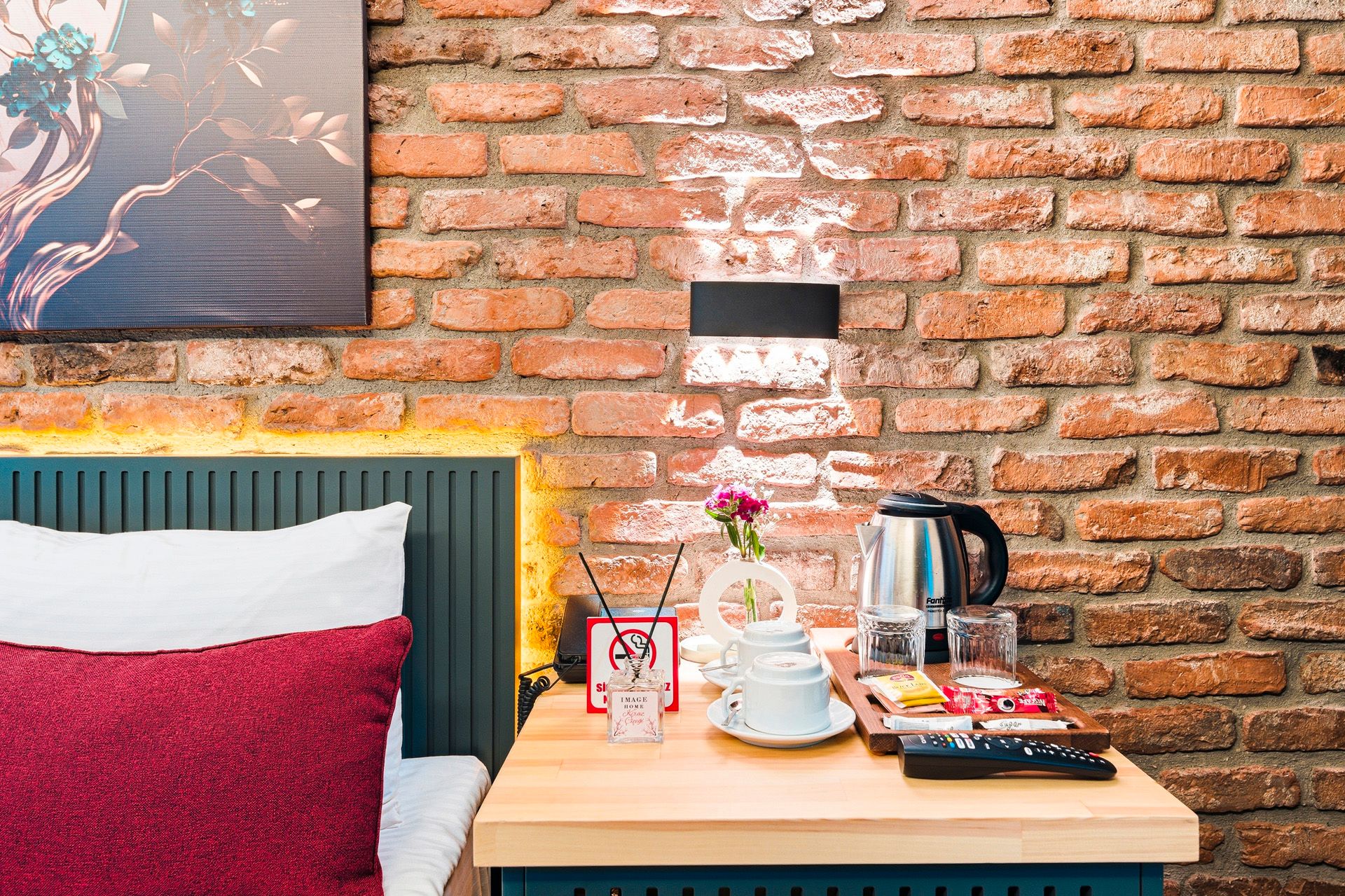 ARTELIST HOTEL | Cozy bedroom nightstand with lamp, flowers, kettle, and snack tray against a brick wall.