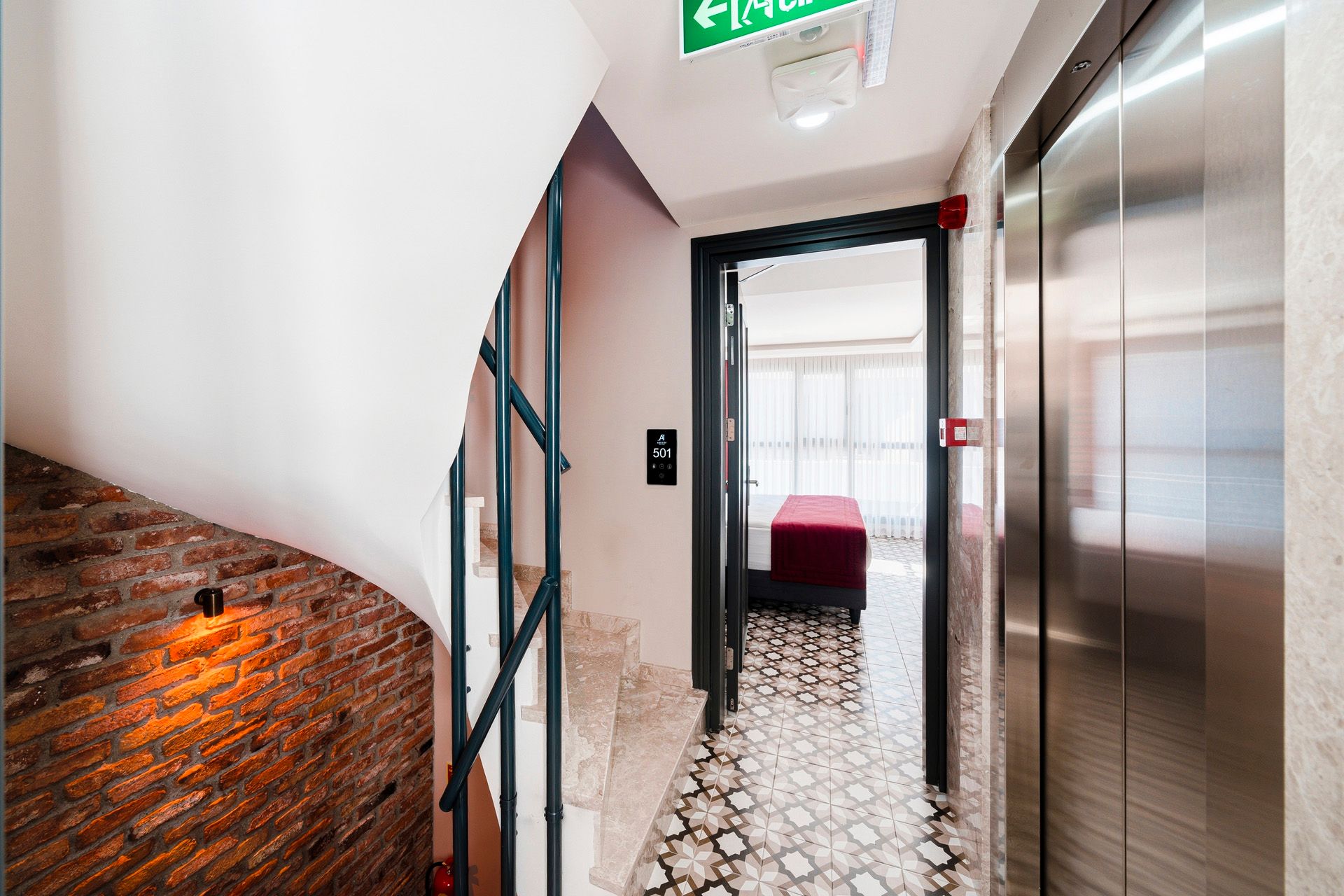 ARTELIST HOTEL | Modern hallway with brick wall, patterned floor, elevator, and a red chair by a window.