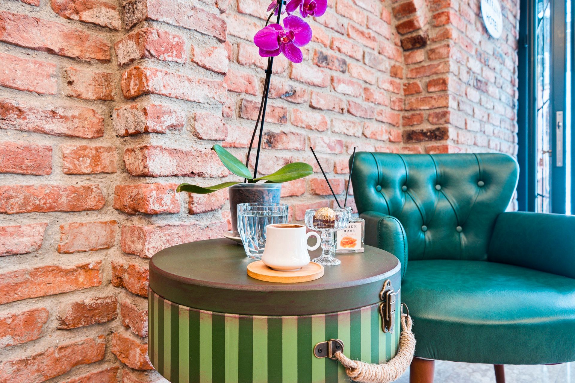 ARTELIST HOTEL | Teal chair beside a striped round table with coffee and purple orchid against a brick wall