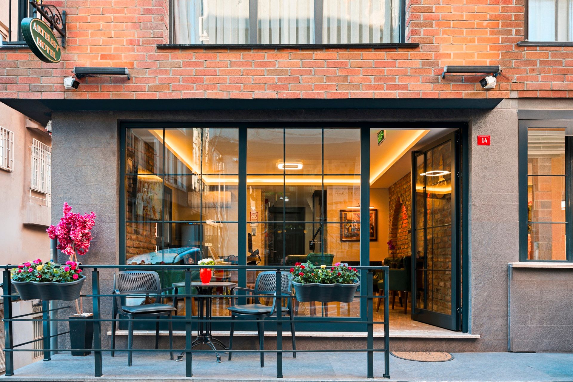 ARTELIST HOTEL | Street-level cafe storefront with large glass windows, patio tables, and pink flowers against a brick facade
