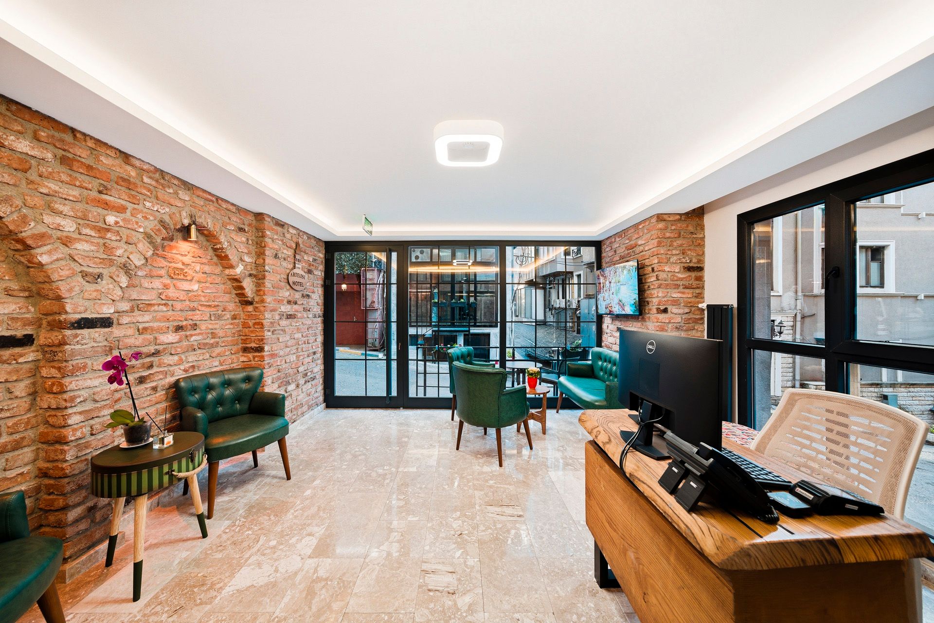 ARTELIST HOTEL | Modern lounge with brick walls, green chairs, wooden desk, and glass doors to an outdoor patio