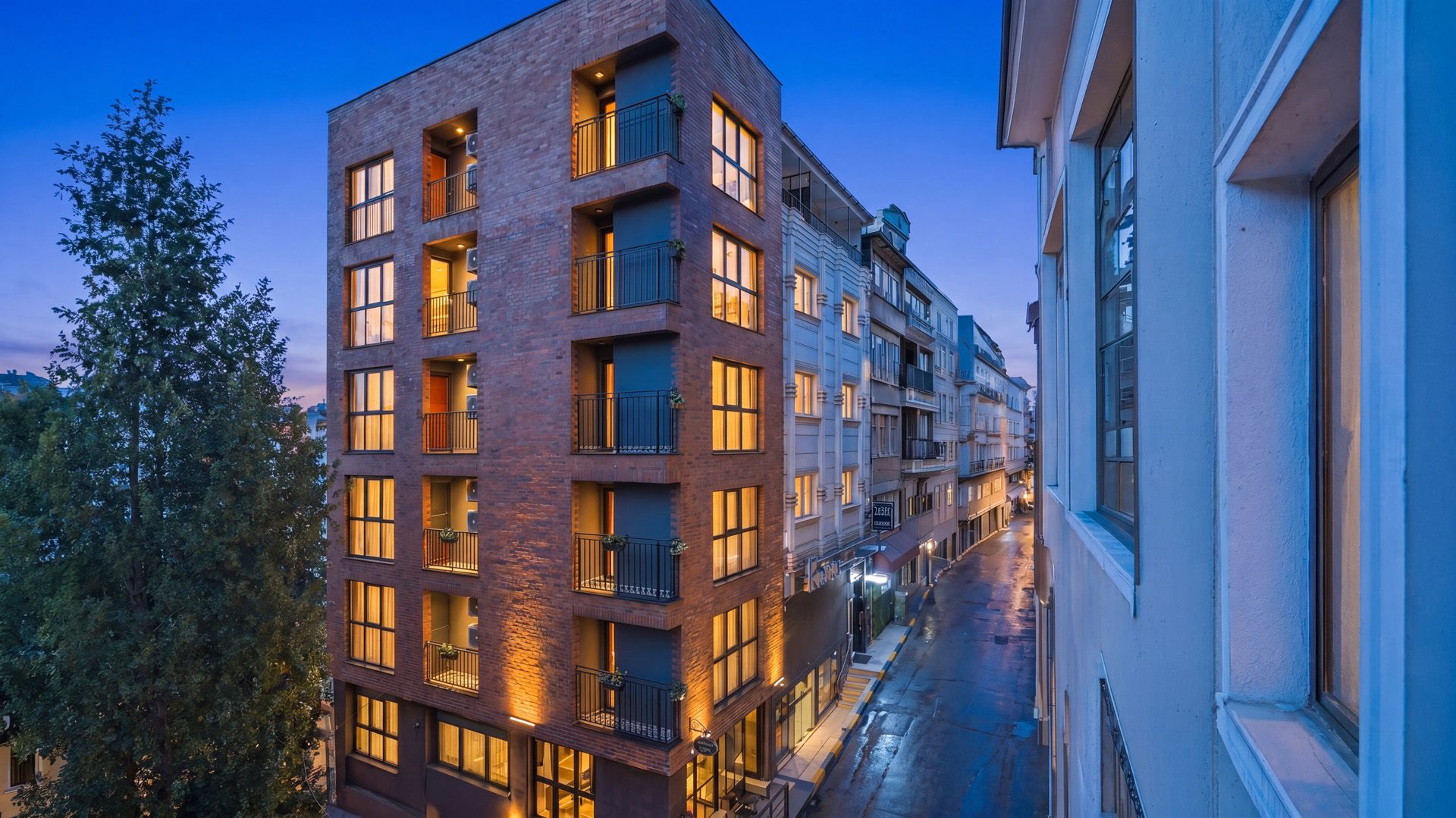 ARTELIST HOTEL | Modern apartment buildings at dusk, with lit windows reflecting on a wet street.