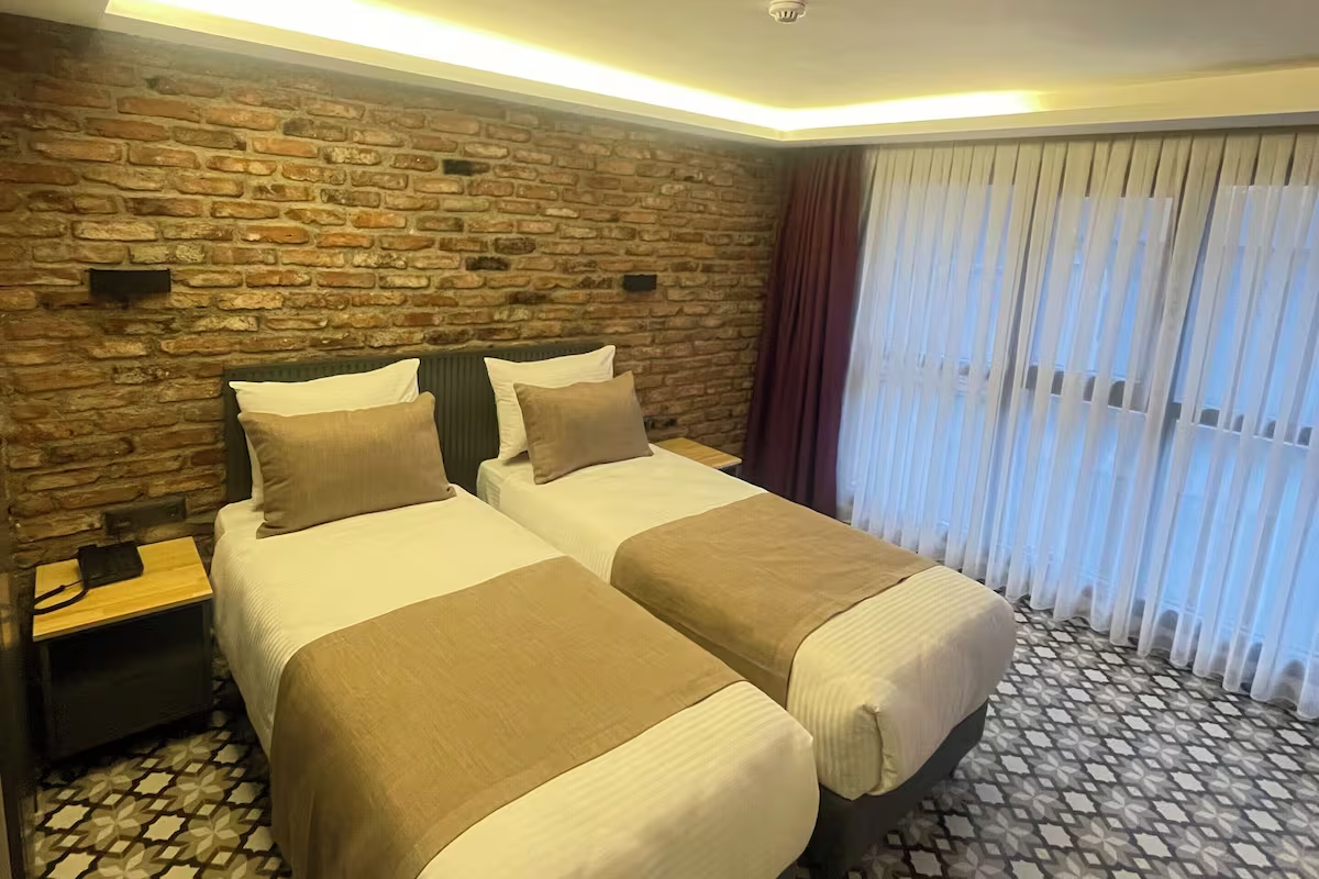 ARTELIST HOTEL | Modern hotel room with two twin beds, brick accent wall, patterned carpet, and large curtained window.