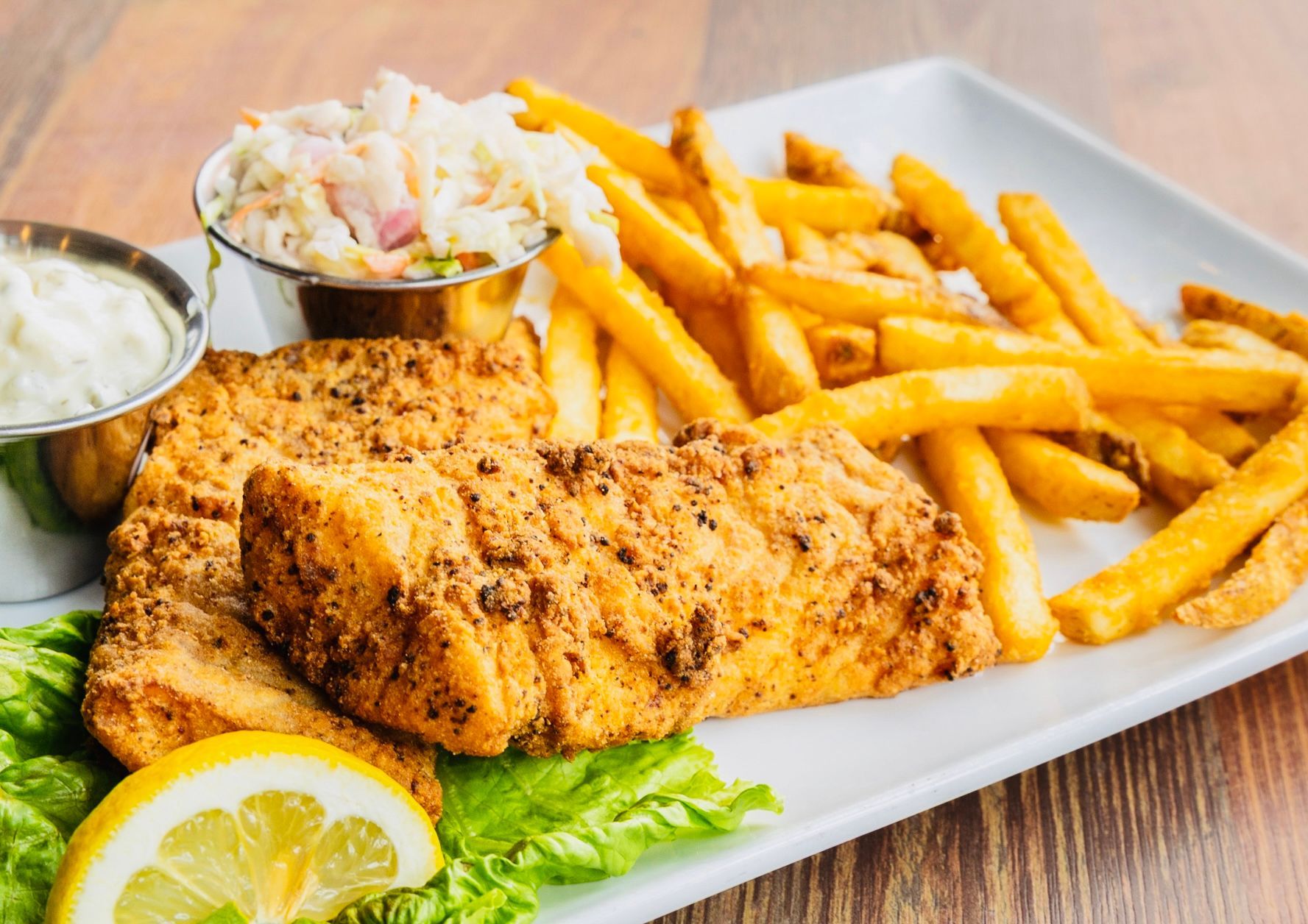Grilled fish fillets with fries, coleslaw, tartar sauce, and lemon wedge on a white plate