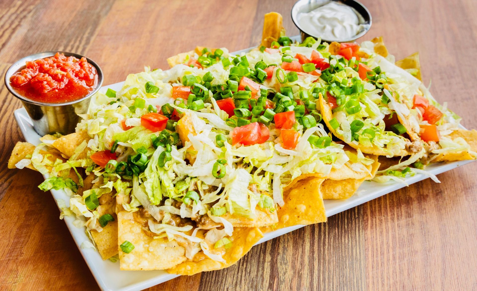 Plate of loaded nachos with cheese, lettuce, tomatoes, and salsa cups on a wooden table