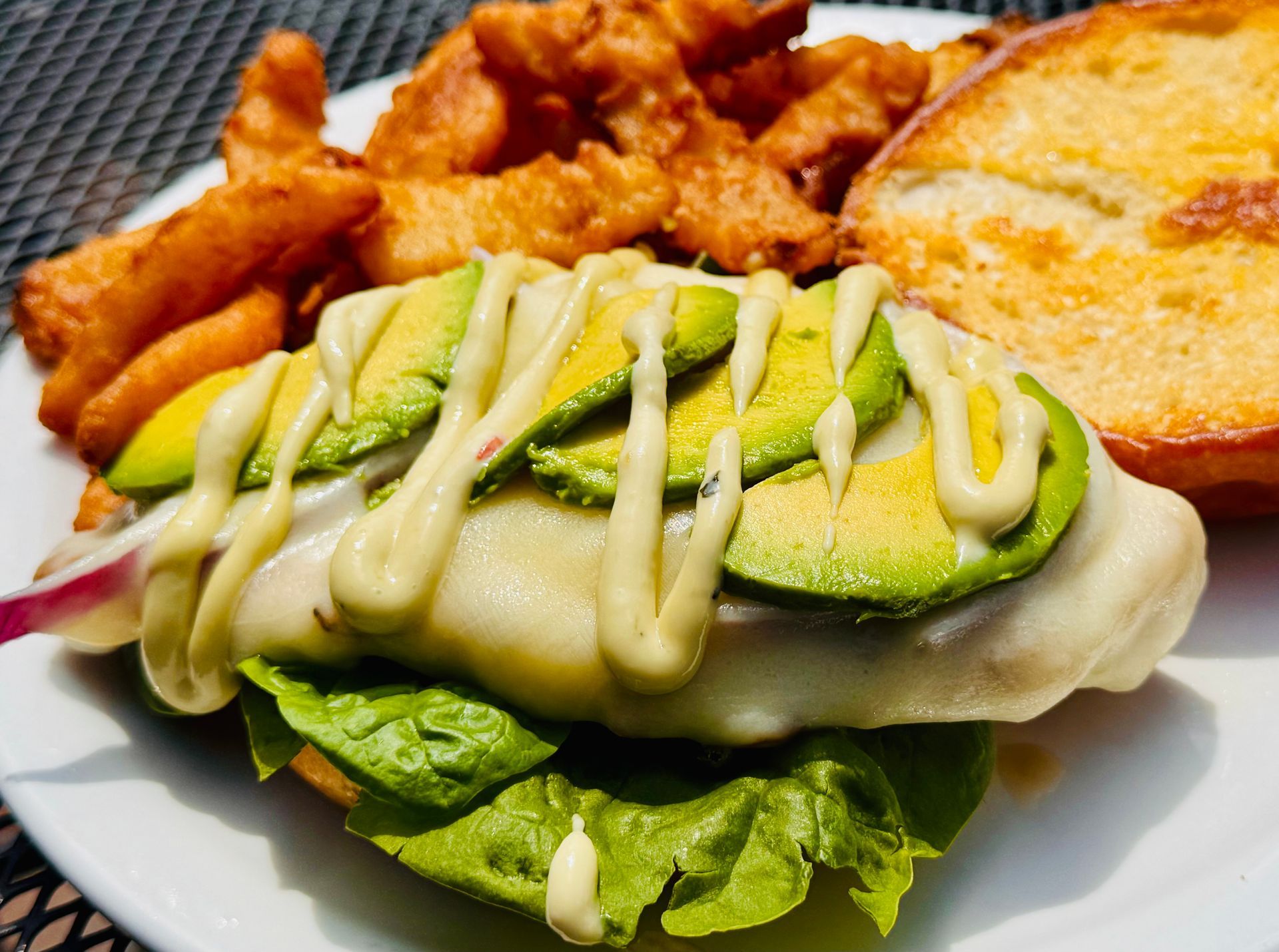 Plated sandwich with avocado slices and creamy sauce beside waffle fries and a breaded side