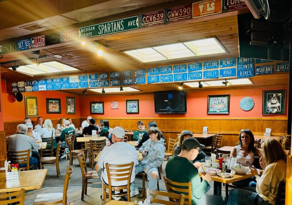 Busy casual restaurant with orange walls, wooden tables, and people dining under colorful menu signs.