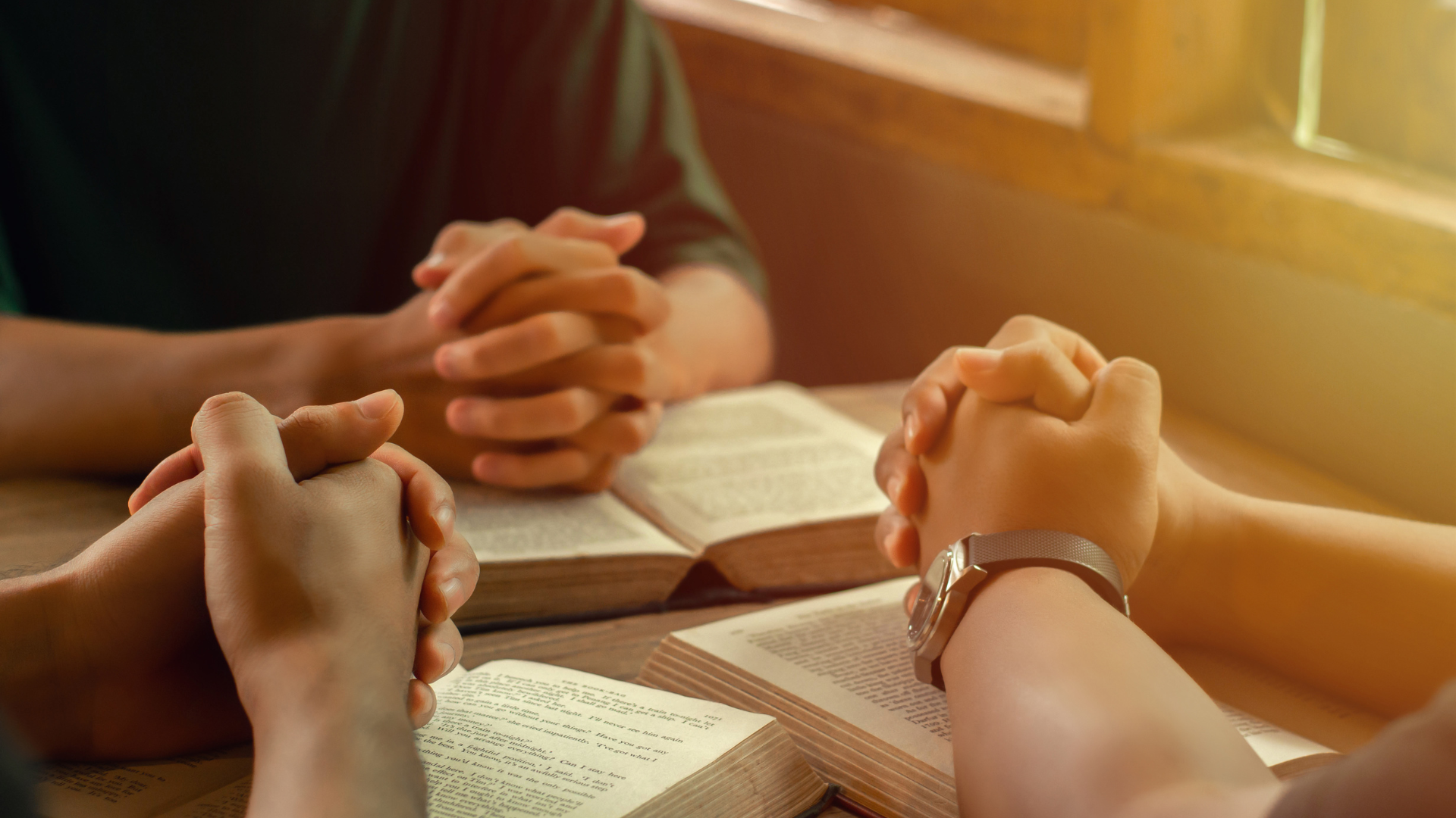 Hands clasped in prayer around an open Bible on a warm-lit table.