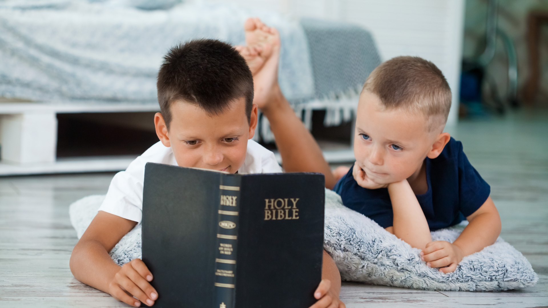 Two children lying on a bed, reading a black Holy Bible together.