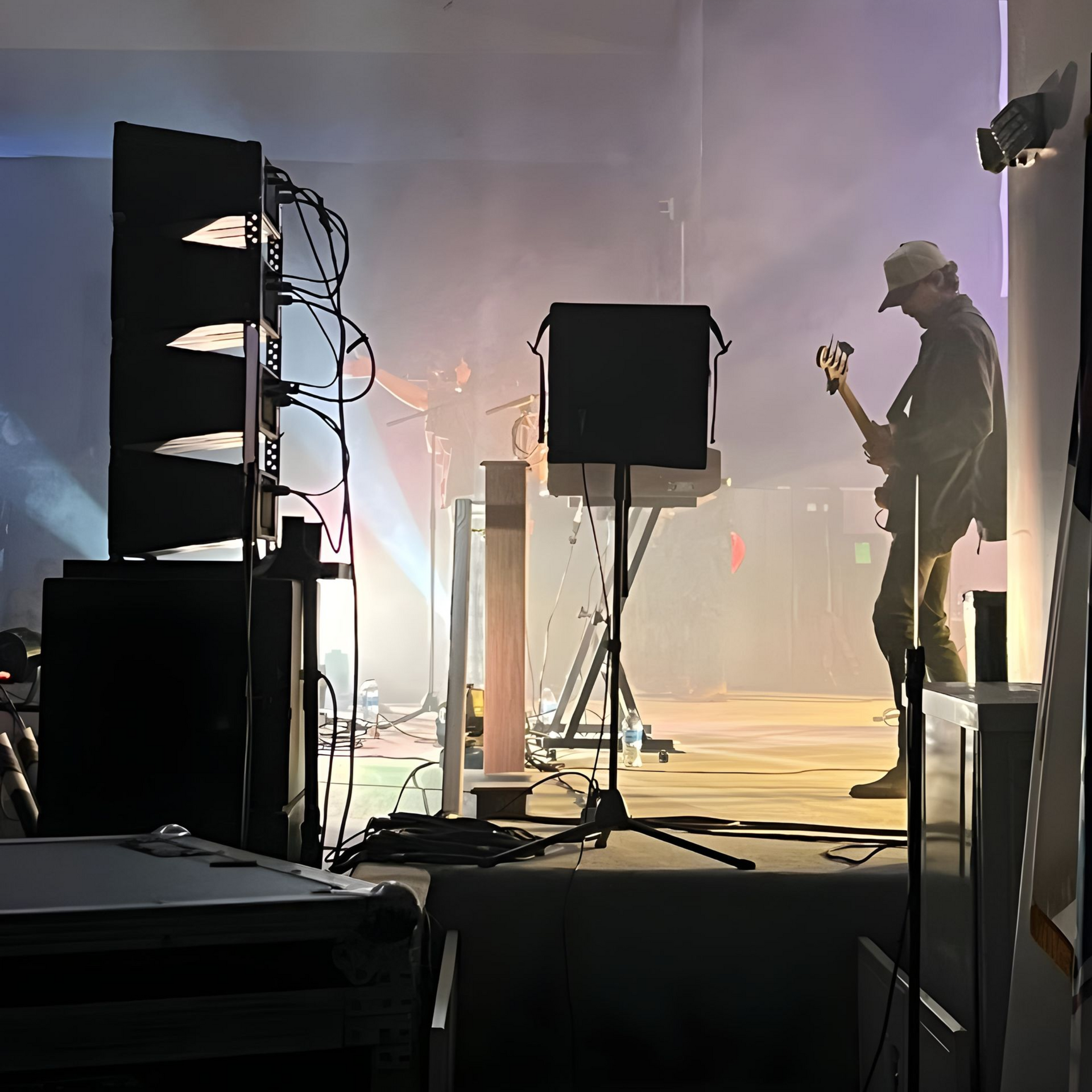 Silhouetted guitarist on a hazy concert stage with speakers, cables, and bright backlighting
