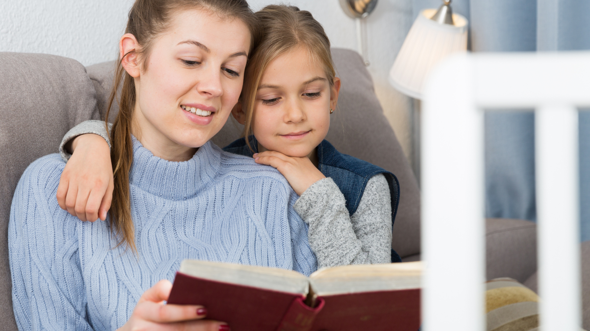 Two people sit on a couch reading a book together, with one smiling and the other leaning in.