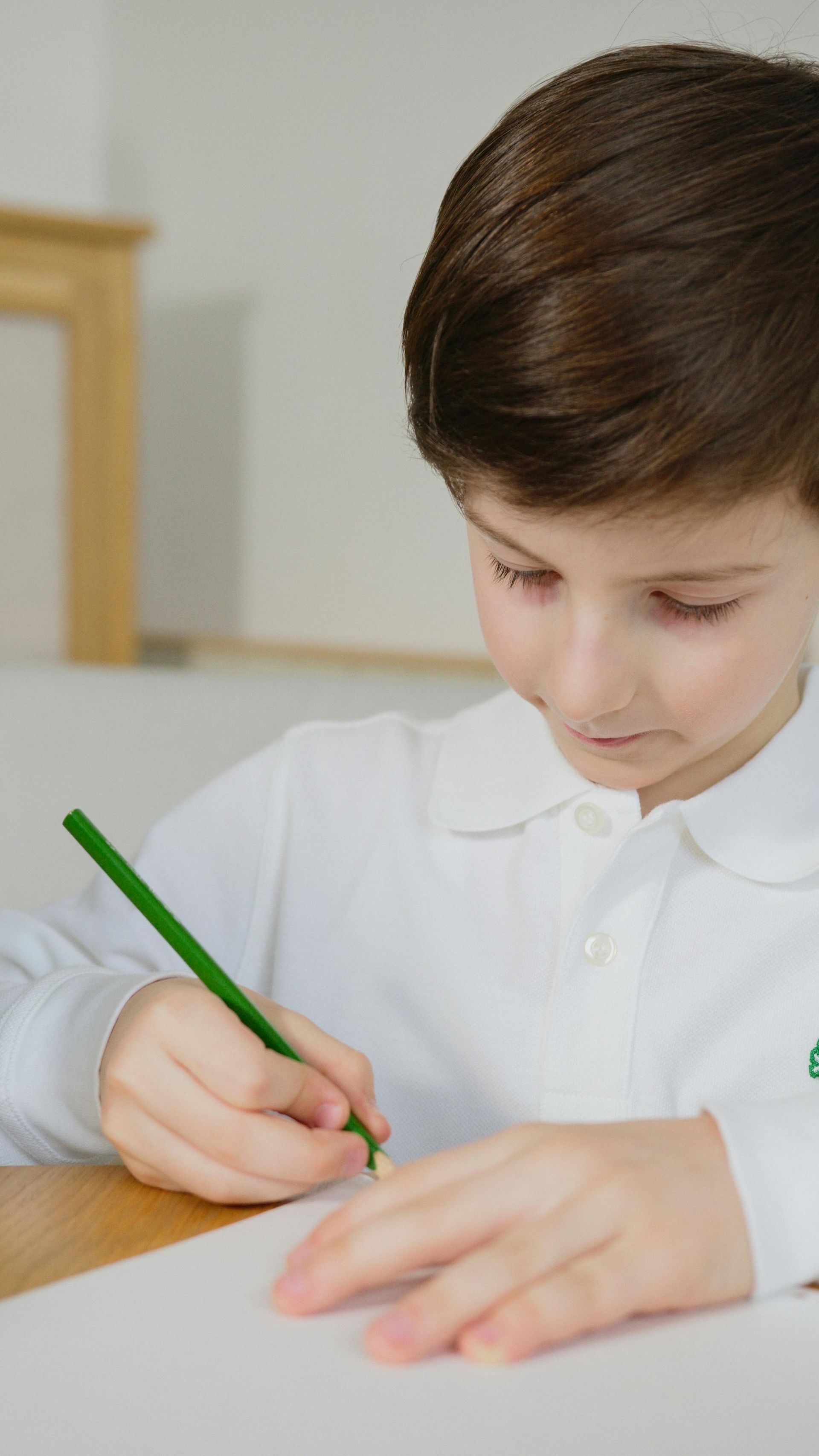 Child writing with a green pencil at a desk in a bright classroom