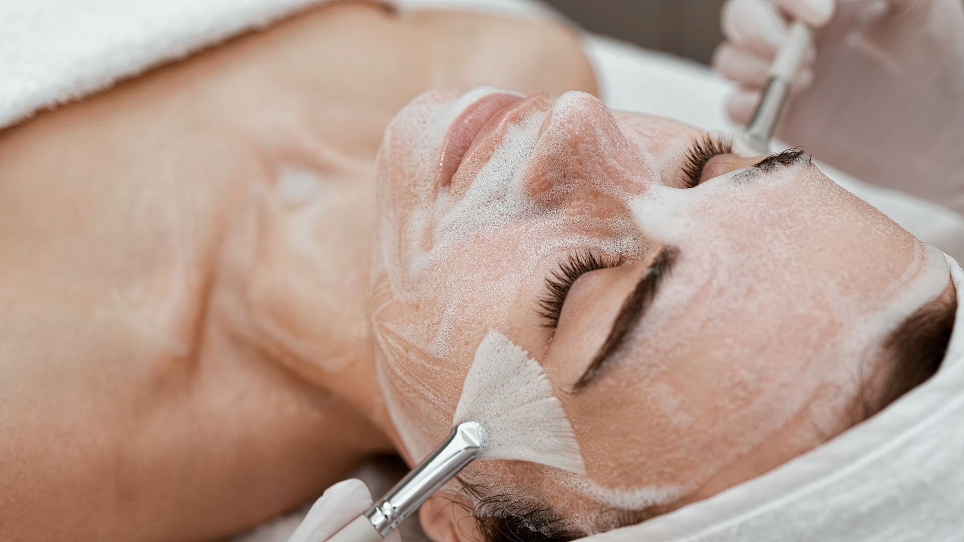 Woman receiving a facial treatment with a brush and cream on her face in a spa setting