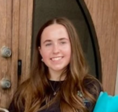 Smiling person standing by a wooden door, wearing a dark top and necklace.