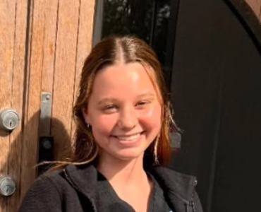 Smiling person in a black top standing by a wooden door in warm sunlight