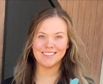Smiling person standing by a wooden door, wearing a black top.