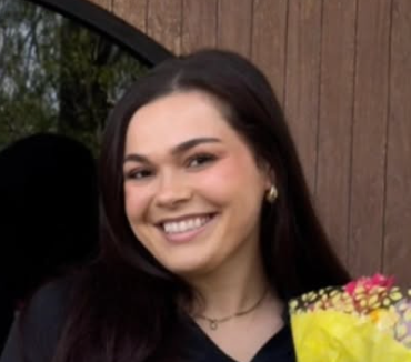 Smiling person with dark hair holding a yellow bouquet outdoors against a wooden wall.