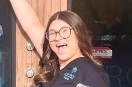 Woman in glasses smiling with arm raised in front of a wooden door