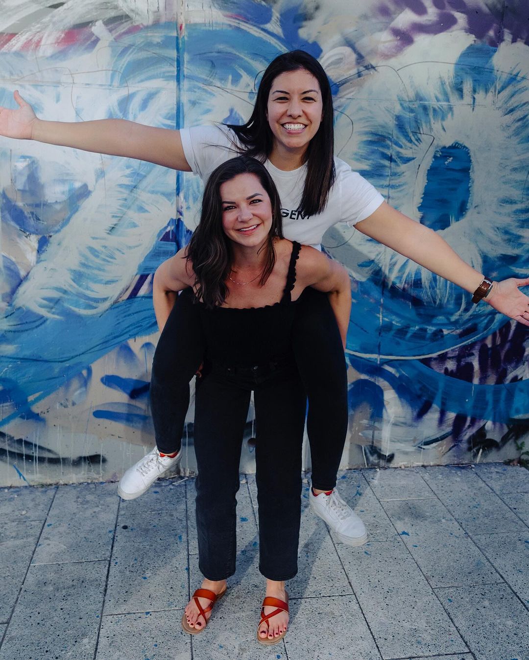 Two smiling friends posing in front of a colorful graffiti wall, one giving the other a piggyback ride.