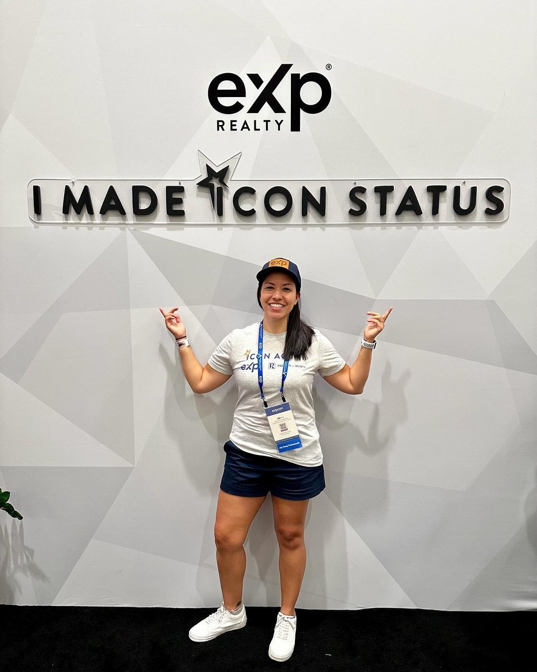 Person posing in front of an eXp Realty wall sign reading “I MADE ICON STATUS,” smiling and pointing upward.