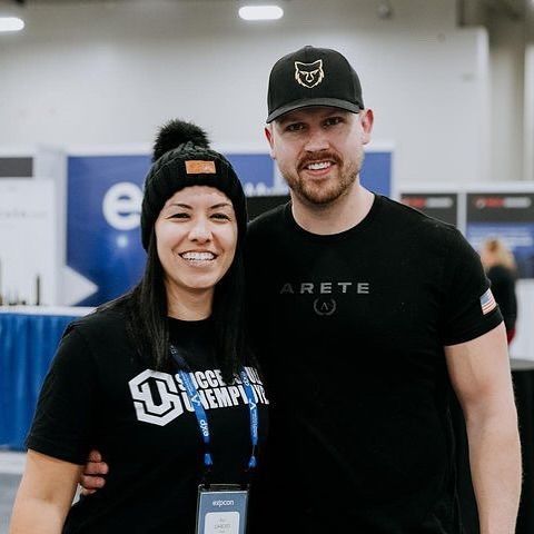 Two smiling people in black shirts and hats standing together indoors at a booth