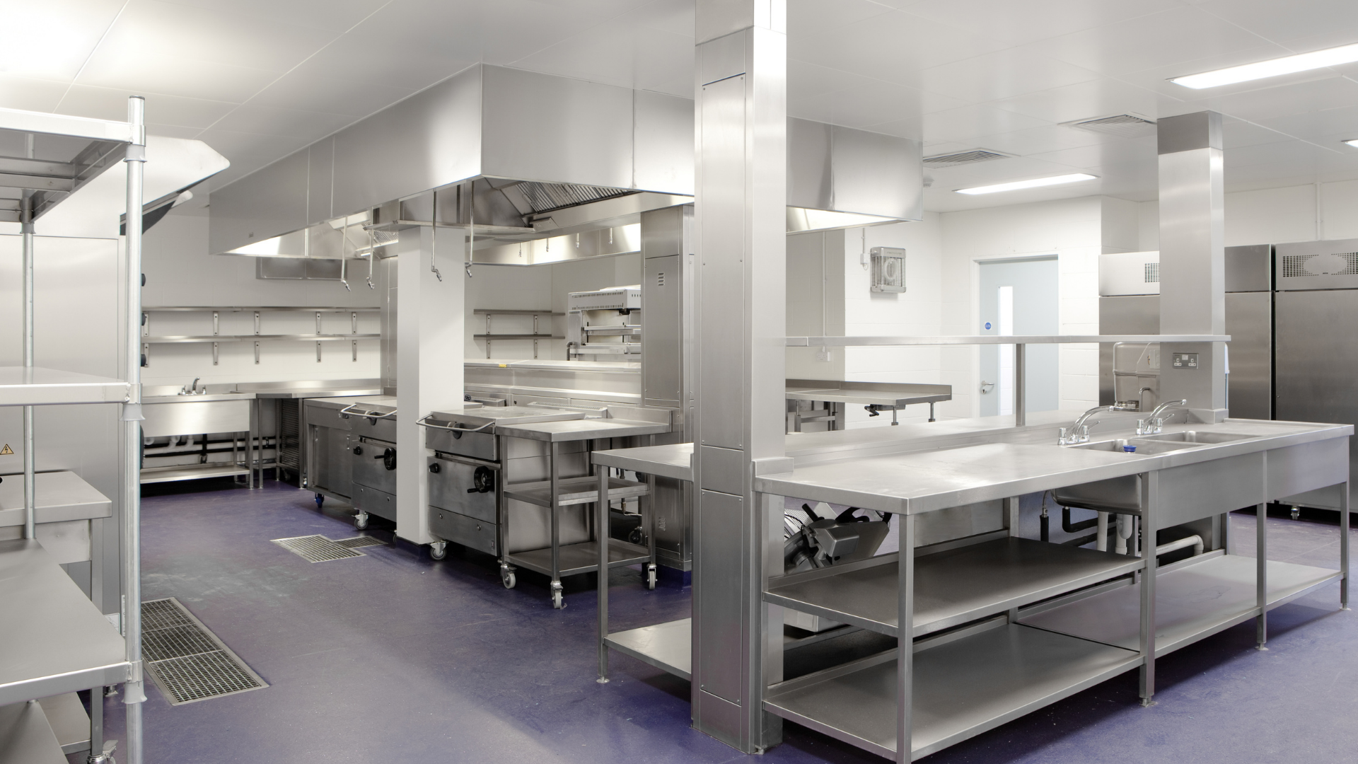 Bright commercial kitchen with stainless steel counters, shelves, and overhead ventilation under fluorescent lights