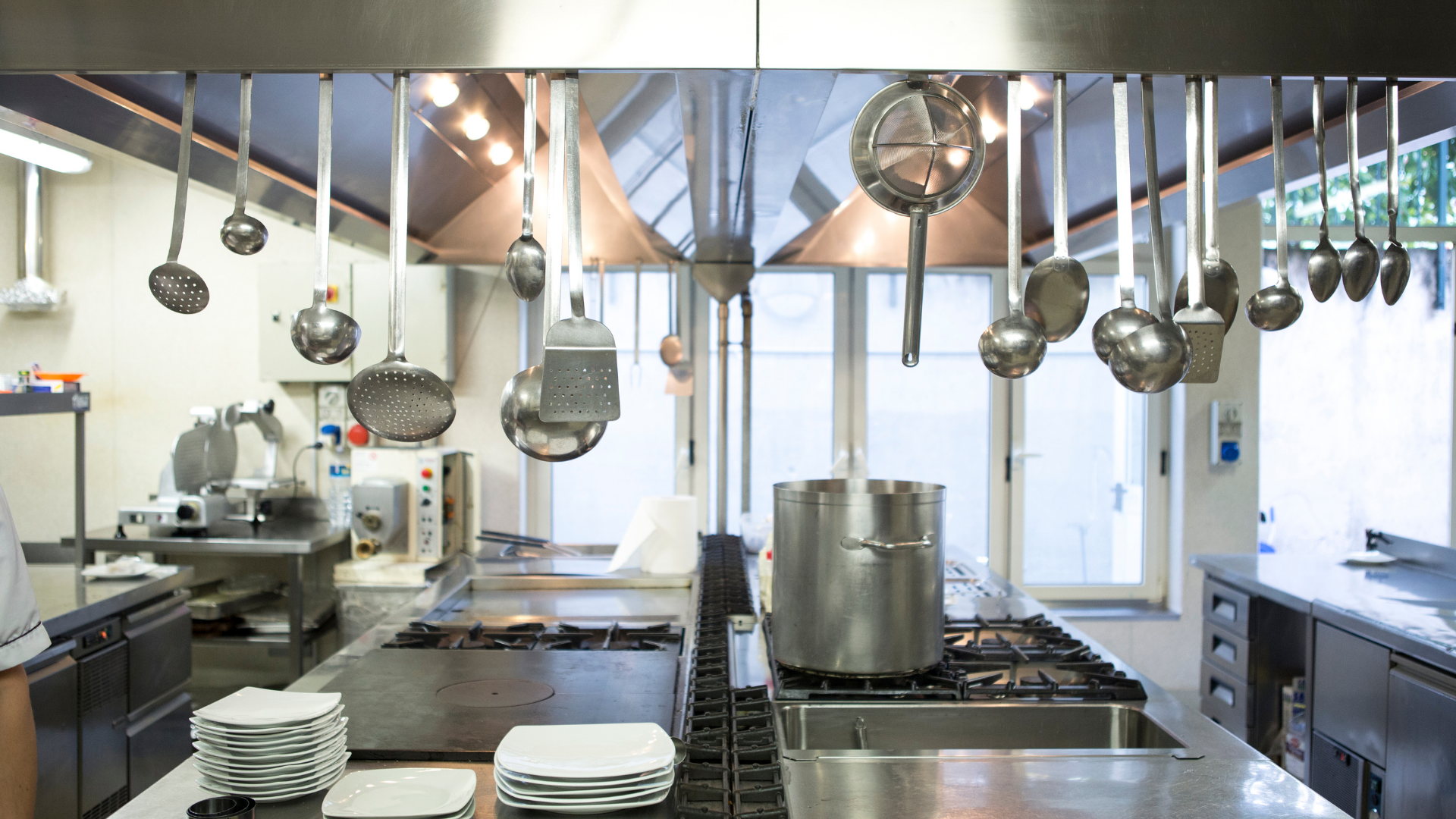 Commercial kitchen with hanging pots, stainless steel counters, stove, and large stockpot by windows