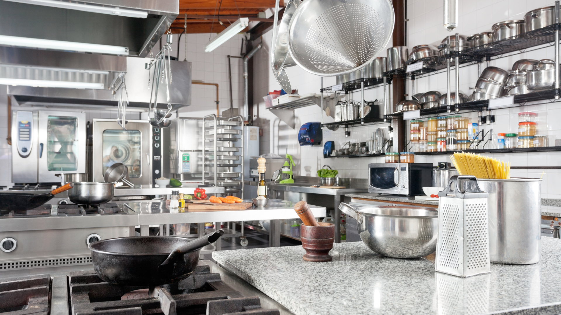 Commercial kitchen with stainless steel counters, hanging pans, and cooking utensils on a prep surface.