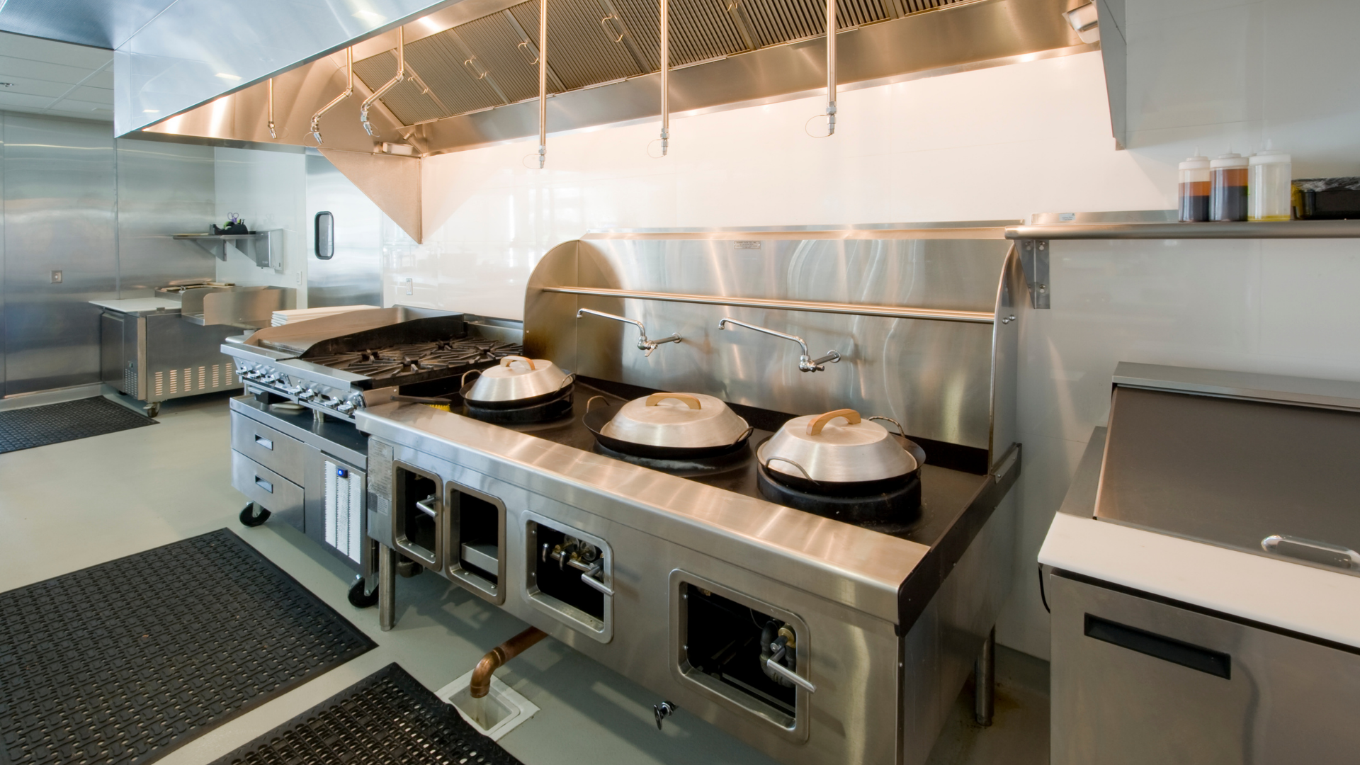Modern stainless-steel restaurant kitchen with covered pans on a cooking line under a large hood