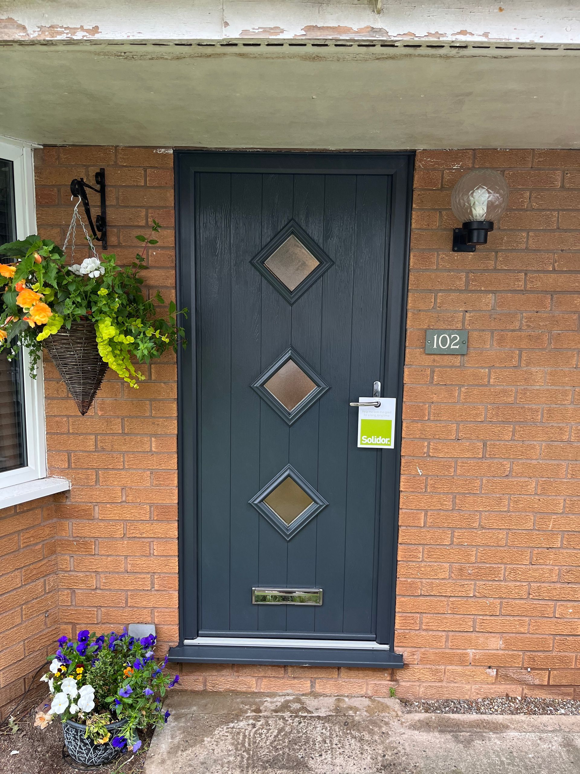 The front door of a brick house with a black door and flowers in front of it.