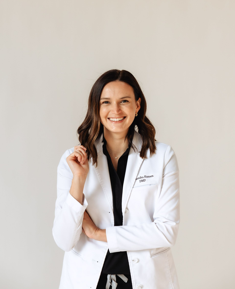 Woman in a white lab coat smiles outdoors; trees in the background.